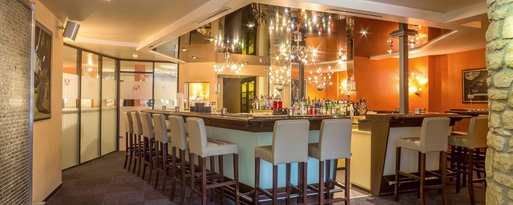 Hotelbar im Hotel Amaris mit modernem Bartresen, Barhockern, großer Getränkeauswahl und stimmungsvoller Beleuchtung.