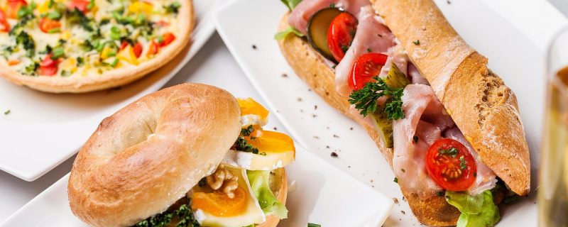 Frisch belegte Sandwiches und Bagel mit Schinken, Käse, Gemüse und Kräutern, serviert auf weißen Tellern.