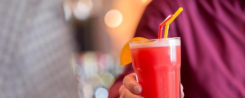 Barkeeper hält einen roten Cocktail in der Hand mit zwei Strohhalmen und einer Orangenschreibe als Dekoration.