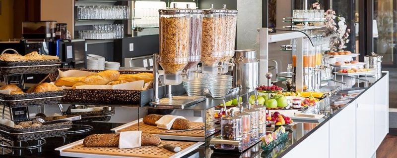 Frühstücksbuffet mit frischen Brötchen, Müsli-Spendern und Obst im modernen Hotelrestaurant.