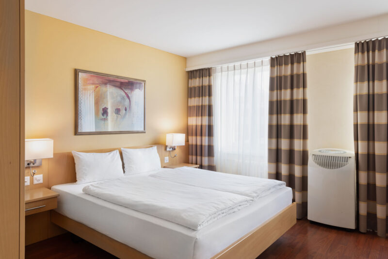 Helles Standardzimmer im Hotel Amaris mit Doppelbett, Nachttischen, Wandbild und großem Fenster mit Vorhängen.