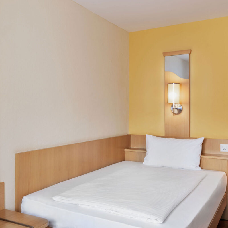 Einzelzimmer im Hotel Amaris mit Einzelbett, Nachttisch, Wandlampe und schlichtem, hellem Raumdesign.