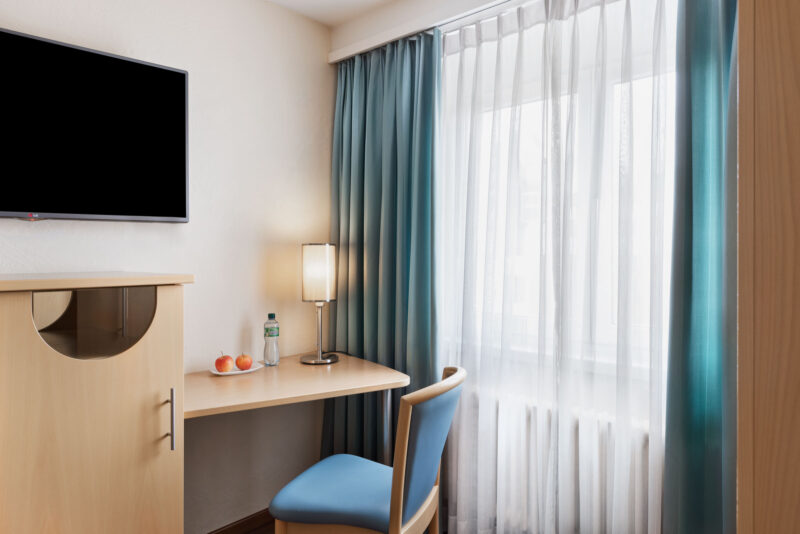 Modernes Hotelzimmer mit Schreibtisch, Stuhl, TV und Tageslicht durch große Fensterfront.
