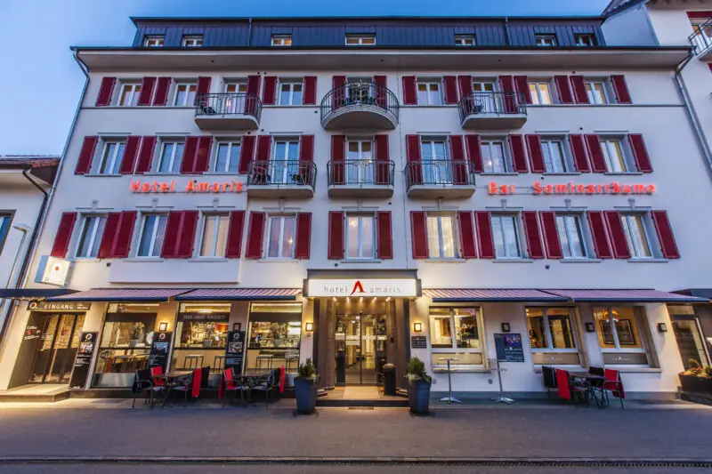 Beleuchtete Fassade des Hotel Amaris mit Balkonen und roten Fensterläden am Abend.